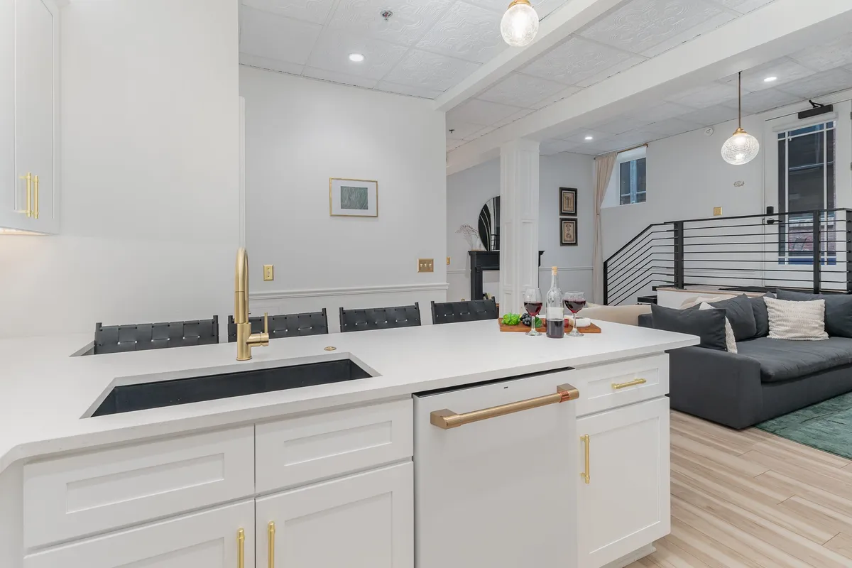 The image presents an open kitchen and living area, featuring a central kitchen island with bar stools, modern sink, sleek appliances, and vibrant artwork in the background. The scene is set for casual entertaining, morning coffee, or pre-evening cocktails—the very activities described in the section.