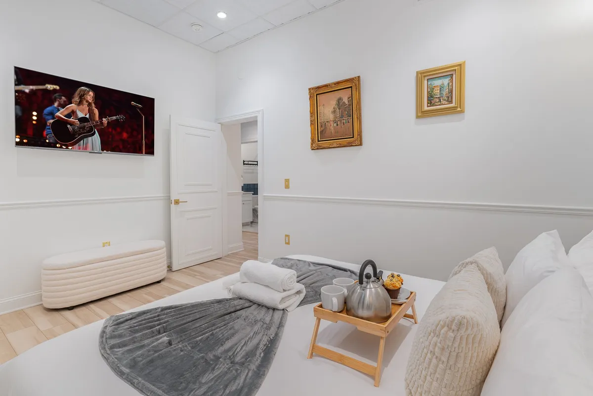 A neatly arranged bed with a tray offering tea and snacks, stylish wall art, and a television displaying a live performance. The space feels cozy, inviting, and tailored for personal downtime or family relaxation.