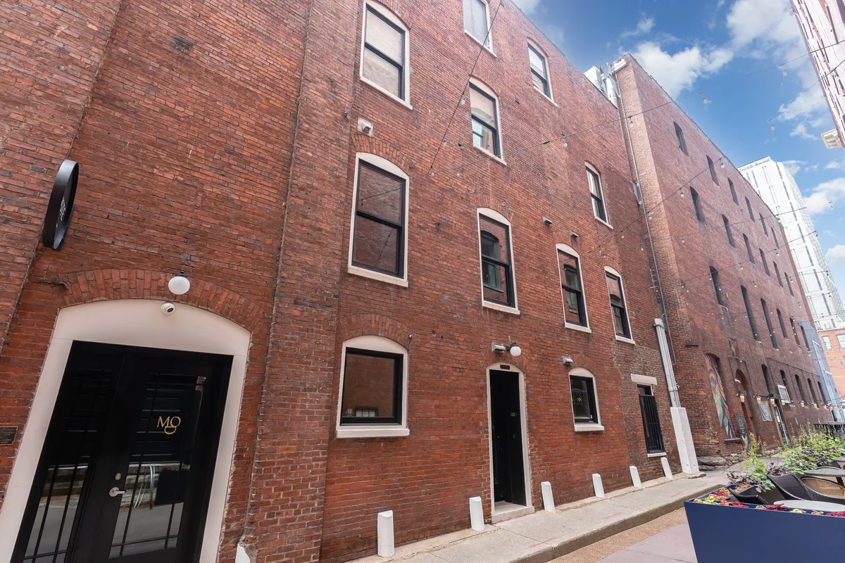 The image presents a classic brick building entrance framed by lush greenery, inviting visitors to explore the vibrant neighborhood. The nostalgic yet lively street scene hints at the charm and variety of local shops and dining options accessible to travelers.