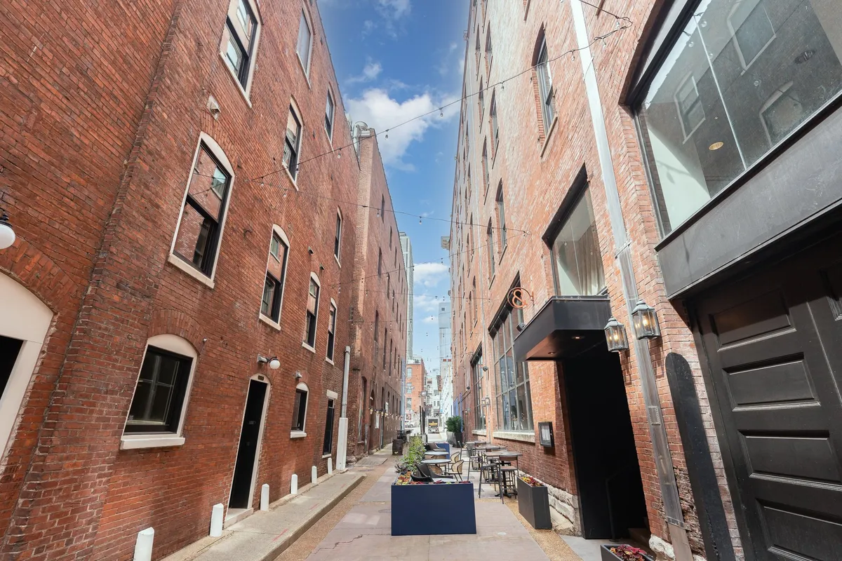A cozy, outdoor seating area between red brick buildings with string lights overhead, creating an inviting alleyway ambiance ideal for evening strolls or relaxed socializing.