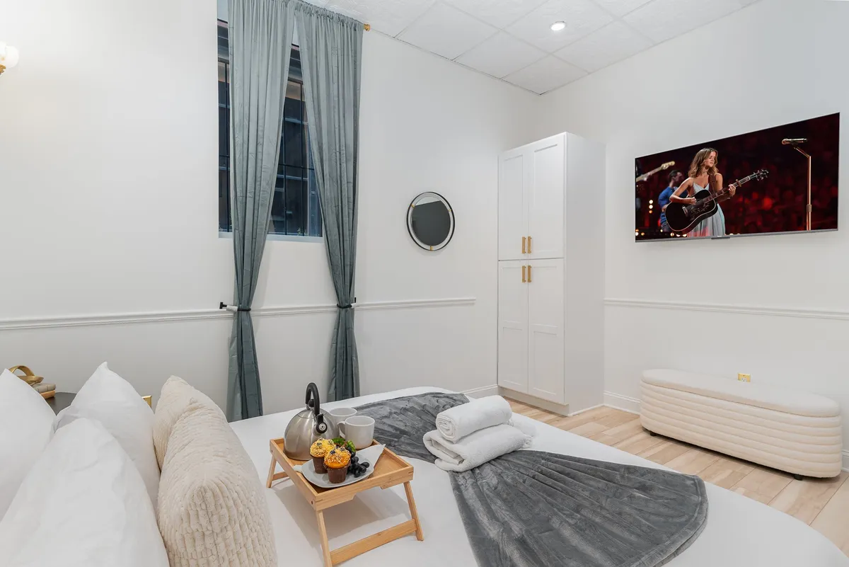 A cozy bedroom scene featuring a bed adorned with decorative pillows, a serving tray with tea and pastries, and a wall-mounted Smart TV set for entertainment. The inviting setup suggests indulgence, whether for a peaceful morning or a cozy movie night.