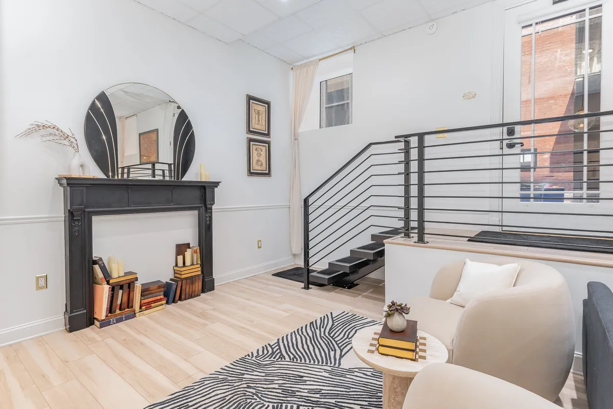 A spacious and inviting interior living area featuring a mirror above a decorative fireplace, a modern staircase, a comfortable seating area, and thoughtfully curated artwork. The space is elegantly styled, evoking a sense of warmth and comfort ideal for socializing or relaxing.