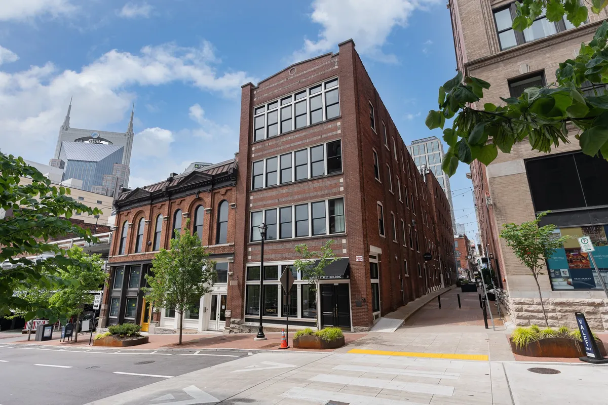 A charming neighborhood streetscape featuring a historic brick building, sidewalk greenery, and glimpses of the city skyline. Cafes and local art spots signal a walkable, lively atmosphere ideal for evening strolls.