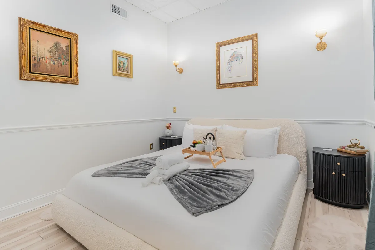 A tranquil bedroom setting featuring plush bedding, decorative pillows, a nightstand with accessories, and elegant wall art. The abundance of natural light and carefully chosen details enhance the feeling of restful comfort.